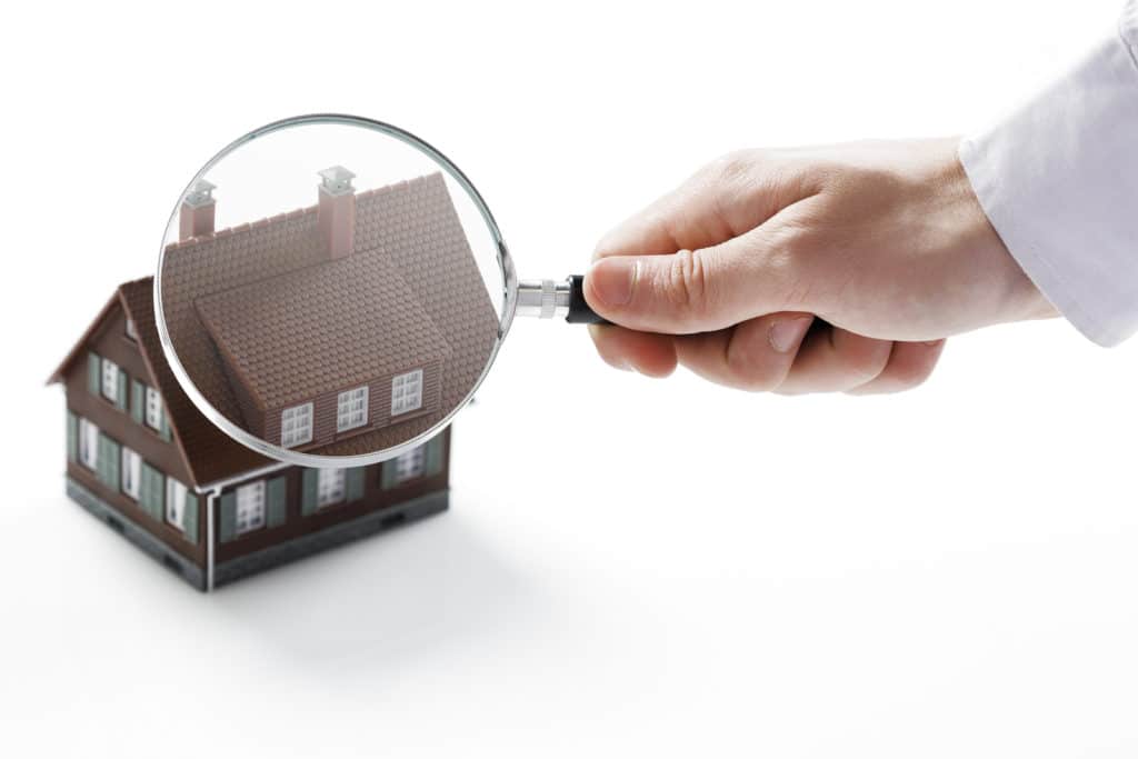 A hand holds a magnifying glass over a miniature house.